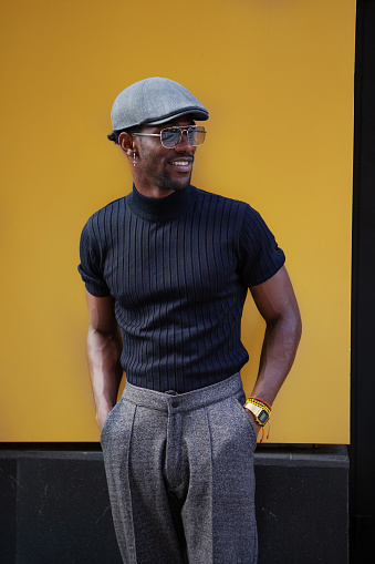 Handsome stylish African American man stands on background of yellow wall outdoors in fashionable attire, wearing cap, sunglasses, black t-shirt and trousers with arrows, modern male fashion model.