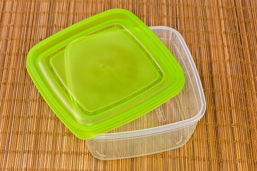 Empty reusable transparent plastic food storage container with pictograms of possible uses on bottom and partly open green lid on a bamboo place mat