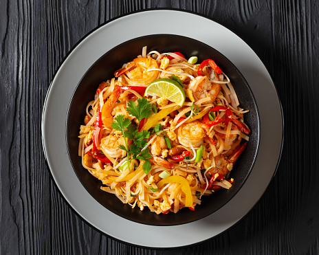 pad thai, phad thai, stir-fried rice noodles with shrimps, peanuts, scrambled eggs, bean sprouts, spring onion, red pepper, lime, in black bowl on black table, free space, flat lay, close-up