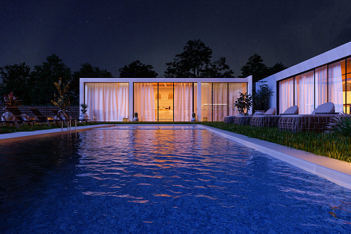 Large swimming pool and patio in the yard of a luxury home at night with a woman standing on a patio in the background
