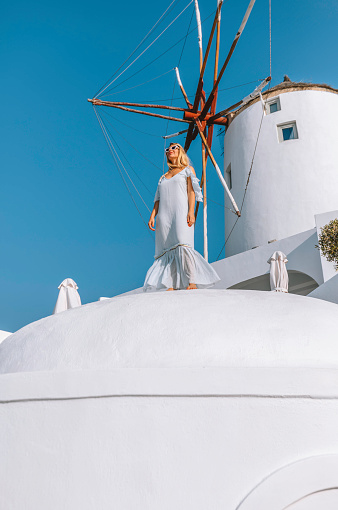 Young beautiful stylish happy female model tourist lady walking and standing on a white washed wall enjoying looking at view the summer day view in a blue color long dress looking at the view of famous traditional old white windmill in Oia or Ia old town, Santorini, Greek islands, Cyclades, Greece at sunset. Travel Greece vacation luxury Europe cruise destination concept