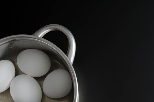 Stock photo showing close-up view of four fried eggs that are being cooked in a stainless steel, non-stick frying pan on a ceramic hob. The pan has rings to crack eggs into in order to keep them separate and stop the egg whites from merging.