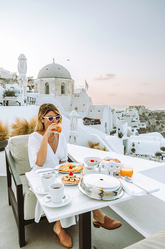A young tourist woman with a white dress enjoys a healthy breakfast on a luxury resort terrace enjoying egg pancakes, and drinking orange juice, tea, and coffee while taking in the stunning Aegean sea view of Oia or Ia Town, Santorini, Greek islands, Cyclades, and Greece.