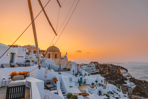The panoramic view of the old town of Oia or Ia on the island of Santorini, Greece, features, white washed houses, blue dome churches, and traditional windmills. The picturesque landscape captures the beauty of the picturesque sun setting in the background with reflection on the ocean water in la Village, a famous spot on the Greek island of Santorini town. The colorful buildings on the cliff side add to the picturesque atmosphere in Cyclades, Greece.