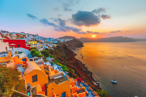 The panoramic view of the old town of Oia or Ia on the island of Santorini, Greece, features, white washed houses, blue dome churches, and traditional windmills. The picturesque landscape captures the beauty of the picturesque sun setting in the background with reflection on the ocean water in la Village, a famous spot on the Greek island of Santorini town. The colorful buildings on the cliff side add to the picturesque atmosphere in Cyclades, Greece.