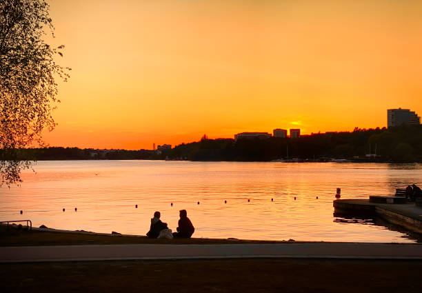 Stockholm, Sweden - May 22, 2022: Beautiful spring city sunset with two people sitting by waters edge with orange sky in Stockholm Sweden May 22, 2022.