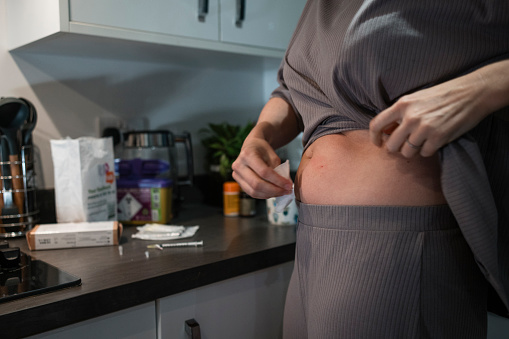 A low angle view of a married lesbian woman who os going through fertility treatment with support from her wife. She is using an alcohol wipe to prep her skin for her hormone therapy injection. On the beach is her medication along with vitamins which she is also taking. She is in the kitchen of her home in the North East of England.