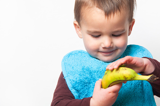 Horizontal medium shot of a child with a blue bib trying to peel a banana to eat it. White background. Isolated.