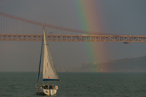 Rainbow over red bridge 25 de Abril Bridge with sailing boat during sunset, Lisbon, Portugal