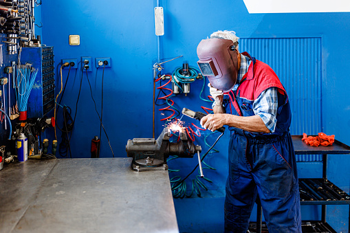 Photo of an Older Professional Worker with Protective Equipment Welding Car Parts due to a Failure.