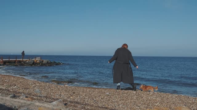 Beautiful redhead Ukrainian woman in coat walks along the sea with small longhaired chihuahua dog in autumn. Slow motion.