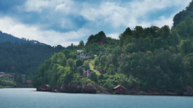 Miniaturized tidy red wooden cabins and boathouses scattered along the forest-covered fjord coast.