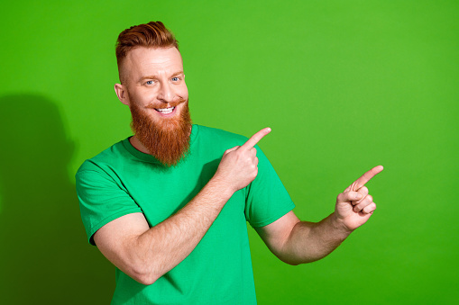Photo of cheerful man with ginger long beard dressed stylish t-shirt indicating at discount empty space isolated on green color background.