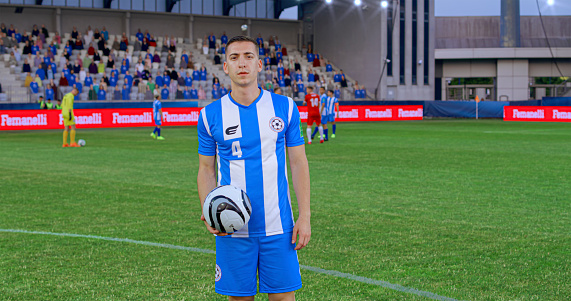 Portrait of male football player in blue jersey standing with football on pitch during match.