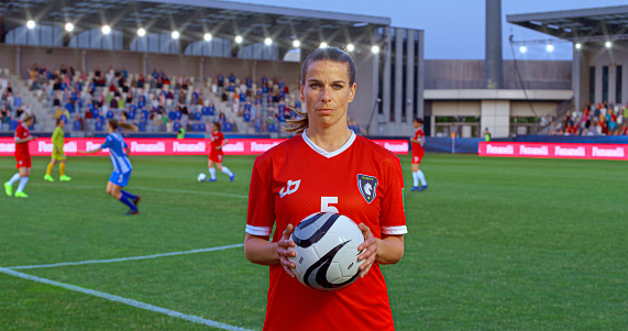 Portrait of female football player in red jersey standing with football on pitch during match.
