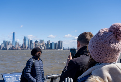 Liberty Island, New York, USA - March, 2024.  Liberty island with tourists in the foreground near the flag pole in front of the Statue of Liberty museum. In the background are the skyscrapers of Wall Street and Lower Manhattan and the Asian tourist is having his photograph taken on a mobile phone with the Manhattan skyline in the background.