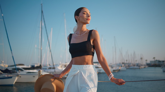 Peaceful woman enjoy ocean pier putting face under evening sunlight. Elegant smiling girl standing on beautiful embankment with luxury yachts. Carefree lady feeling calmness relaxing on sea waterfront