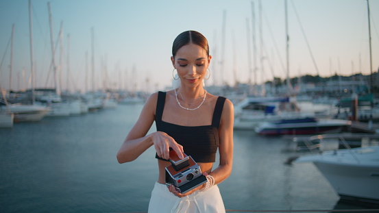 Happy woman adjusting photocamera at beautiful evening pier close up. Relaxed attractive girl taking retro camera to make marine landscape photo. Stylish young lady photographing luxury yachts at quay