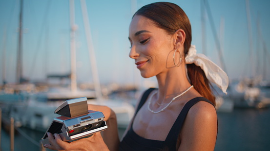 Smiling woman holding photocamera on evening waterfront close up. Carefree young lady fold modern photo camera after taking picture. Elegant happy girl standing on embankment enjoy summer sunlight.