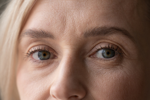 A close-up view captures the top half of a womans face, revealing her striking blue eyes, soft blond eyebrows, and delicate skin texture. The focus on her eyes creates an intimate and introspective mood.