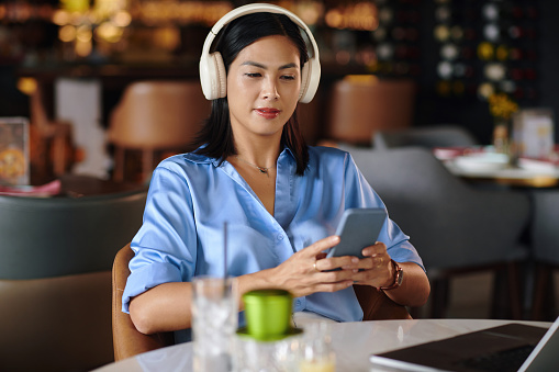 Woman in headphones sitting at restaurant table and listening to music on smartphone