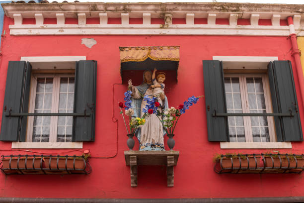 This is an image of a religious icon depicting A Burano con Romeo on a plinth affixed to the wall of a house. There are also two vases containing flowers. The icon is in between two shuttered windows.