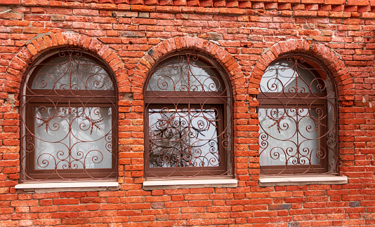 This is an image of Three arched windows in a red brick wall. Each window is protected by decorative metalwork.