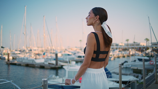 Romantic girl looking yachts moored at pier summer evening. Dreamy beautiful woman posing on quayside looking camera with gentle smile. Stylish tanned tourist contemplating luxury white boats alone.