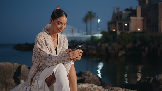 Stylish traveler scrolling smartphone at evening coastal town. Elegant woman looking cellphone screen with smile reading message outdoors. Happy girl browsing mobile phone application on quay street.