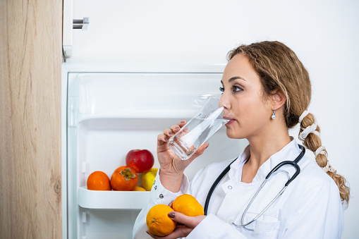 A young female doctor in a gown with a stethoscope stands by the refrigerator and looks at tangerines, drinks water from a transparent glass.