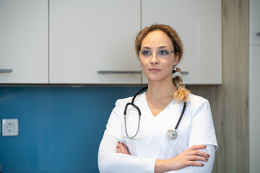 Beautiful smiling intern holds clipboard with written papers and pen, wears white uniform, ready to see patient in own private clinic, stands indoor against white background. Copy space area