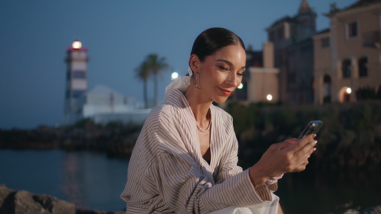 Smiling lady surfing internet smartphone at twilight waterfront close up. Happy relaxed woman watching social media on cellphone sitting evening coastal town. Elegant businesswoman relaxing with coast