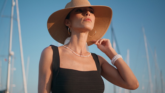 Straw hat girl relaxing on sunny nature close up. Portrait of elegant tanned woman posing in front yachts masts at summer vacation. Trendy fashion model in luxurious headdress standing at quay.