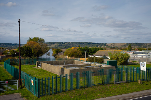 Tidal Sluice, Hythe, Kent - A security fenced-in Grand Redoubt Tidal Sluice  with a drain leading to a horizon of trees and hills on a sunny day, with a caravan park.