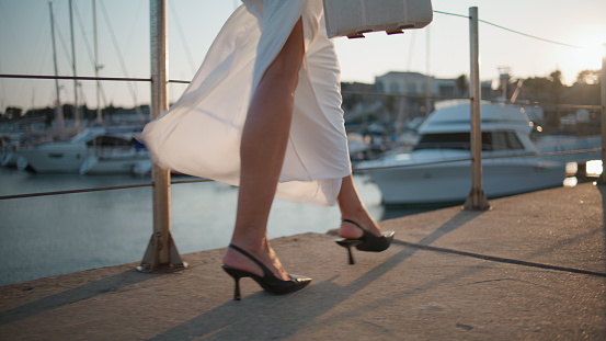 High-heels legs going on embankment summer windy evening close up. Unrecognizable stylish woman walking at waterfront in elegant white clothes. Unknown relaxed girl strolling pier near luxury yachts.