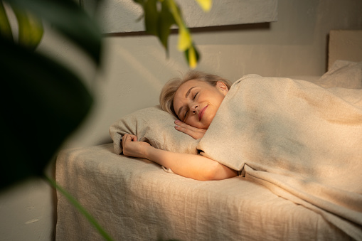 A woman is captured in a serene slumber, nestled under a light blanket in a warmly lit, inviting bedroom setting. The room exudes a tranquil atmosphere, suggesting a late evening or nighttime environment, conducive to rest and relaxation.