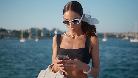 Fashion woman texting mobile phone on beautiful ocean waterfront close up. Carefree trendy girl smiling messaging on smartphone at beach. Luxurious female traveler in sunglasses looking cellphone.