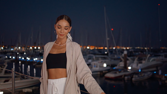 Romantic woman posing at evening embankment in stylish clothes. Carefree relaxed girl looking camera relaxing at night quay with moored luxury yachts. Beautiful trendy brunette standing at waterfront.