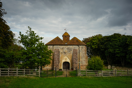 The exterior of St Marys Church in East Guldeford with cemetery, surrounded by trees, with grass in the foreground and a moody sky.