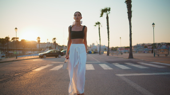 Rich businesswoman walking at sunset road in trendy outfit. Confident brunette woman going on asphalt roadway at summer evening. Trendy successful lady strolling town enjoying beautiful sundown.