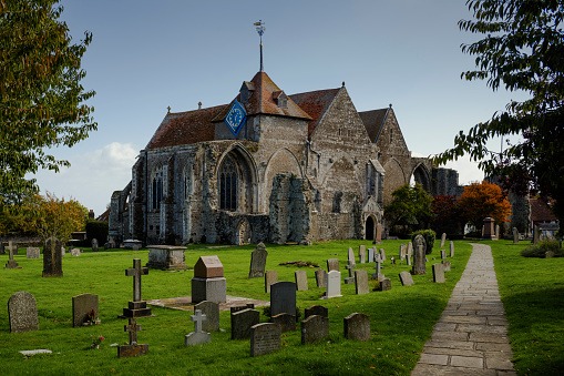 A wide view of the Church of St Thomas in Winchelsea, with gravestones, a pathway, trees and a blue sky.