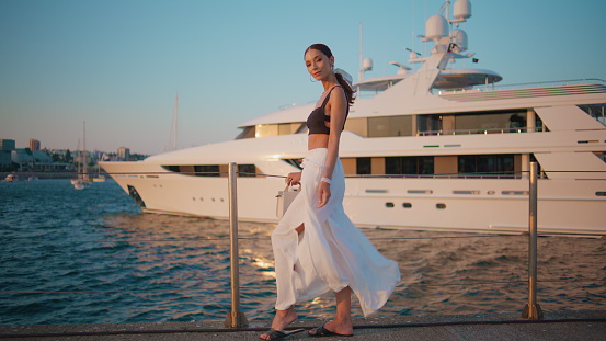 Attractive lady relaxing at quay enjoying windy summer evening. Romantic dreamy woman posing in front luxury white yacht. Elegant young brunette traveler standing on beautiful waterfront at weekend.