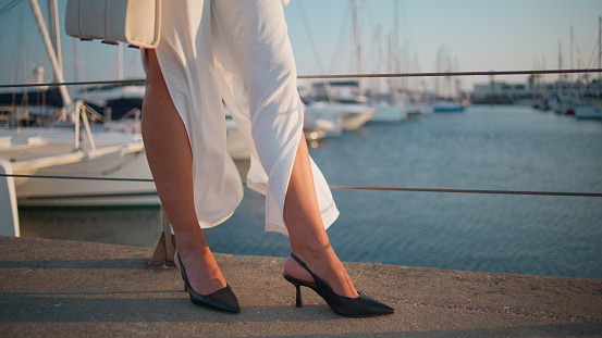 Slender female legs standing at embankment in high-heels summer evening close up. Unrecognizable woman in trendy pants posing at sunny pier. Unknown fashion lady relaxing near luxury yachts on quay.