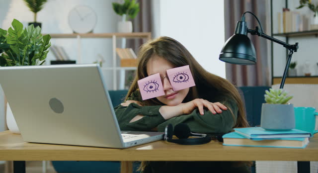 Beautiful tired woman falling asleep on the table near laptop and putting on glasses with funny stickers with drawn open eyes