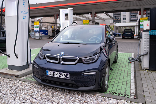 BMW i3 electric car is being charged from an electric charging station, charging cable, another gas station for traditional gasoline cars visible in background, Berlin, Germany - February 17, 2024