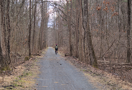 woman riding on a gravel bike path (cycling on unpaved dirt trail) distance, far away, from behind (woods, forest, trees)