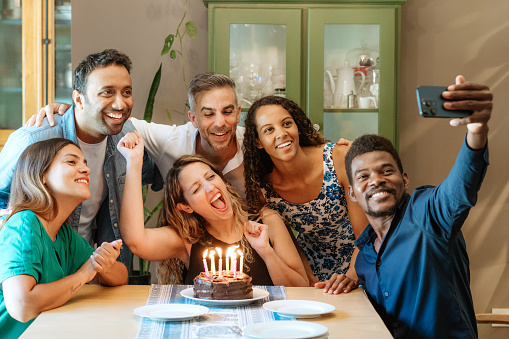 A diverse group of friends gather around a birthday cake, capturing memories with a selfie at a warm, friendly home celebration.