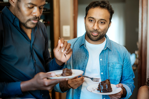 Two male friends sharing a moment of joy while eating chocolate cake at an informal social event.