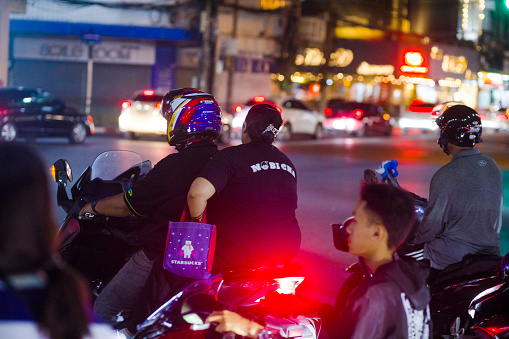 Adult thai people on motorcycles are waiting at road intersection and stoplight at junction Ratchadaphisek Rd and ladprao Rd at night. People are standing in front of queued traffic. Focus is on women in center. In background traffic is ruling on Ladprao Rd.