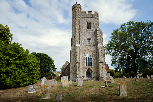 An exterior of St Mary the Virgin Church in, Stone in Oxney, with gravestones and trees, on a sunny day.